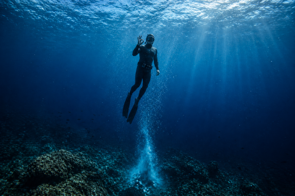 غواص حر في أعماق البحر يشير بعلامة “OK” أثناء الصعود مع آثار فقاعات في الماء الأزرق